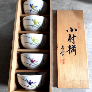 Puede incluir: Un juego de cinco cuencos de cerámica blanca con motivos florales pintados a mano en azul, amarillo y morado, dispuestos en una caja de madera vertical. La tapa de madera presenta caracteres de caligrafía japonesa y sellos rojos.