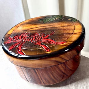 Puede incluir: Un recipiente redondo de madera con tapa y acabado pulido. La tapa presenta un diseño tallado y pintado de una gran flor de crisantemo roja y hojas verdes sobre un fondo de madera natural con bordes de color marrón oscuro.