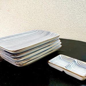 Puede incluir: Una pila de cinco platos rectangulares de cerámica blanca con finas rayas azules horizontales y bordes dorados, acompañada de un plato rectangular más pequeño y separado con dos compartimentos, rayas azules a juego y borde dorado.