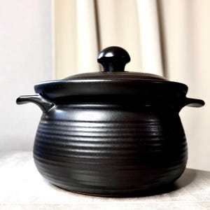 May include: A matte black ceramic pot with a matching lid and two side handles. The body of the pot features a series of horizontal ridges circling the exterior, while the lid is topped with a small, rounded knob handle for easy lifting.