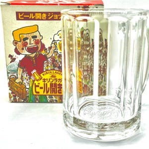 以下が含まれることがあります： 縦の溝と持ち手が付いた透明なガラス製ビールジョッキ。箱にはビールジョッキとスーツケースを持った人物のカラフルな漫画イラストが描かれており、日本語のテキストとキリンラガーのロゴが印刷されています。