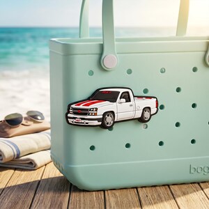 May include: A pale green tote bag with a white pickup truck-shaped charm. The truck has red and white stripes and the text "RST" on the windshield. Sunglasses and a folded towel are visible.