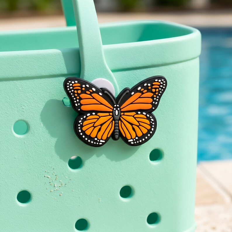 May include: A monarch butterfly charm in black, orange, and white is attached to a light green tote bag. The butterfly has detailed wing patterns and is made of a rubbery material. The bag has a handle and several round holes.