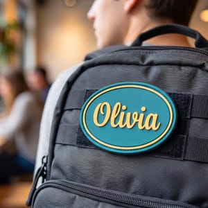 May include: A teal and gold name patch with the name "Olivia" in gold script lettering is attached to a black backpack. The patch is oval-shaped with a gold border.