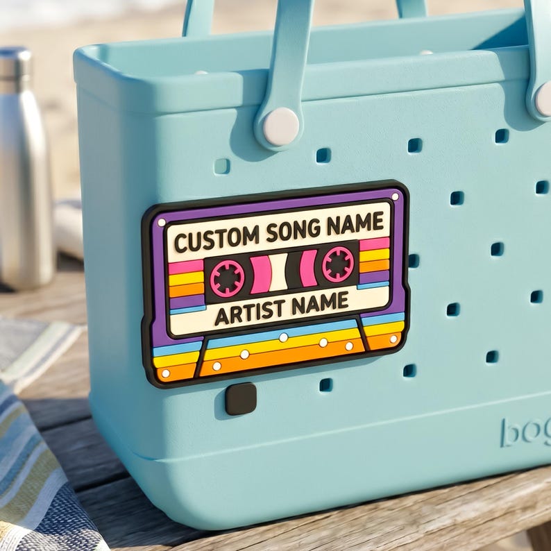 May include: A light blue tote bag with a retro cassette tape design. The cassette tape has a rainbow stripe design and the text "CUSTOM SONG NAME" and "ARTIST NAME". The bag has two handles and a silver water bottle is visible.