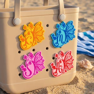 Könnte beinhalten: Beige Tragetasche mit vier schmetterlingsförmigen Anhängern in Gelb, Blau, Pink und Rot. Jeder Anhänger hat ein Buchstabendesign. Die Tasche befindet sich an einem Sandstrand.