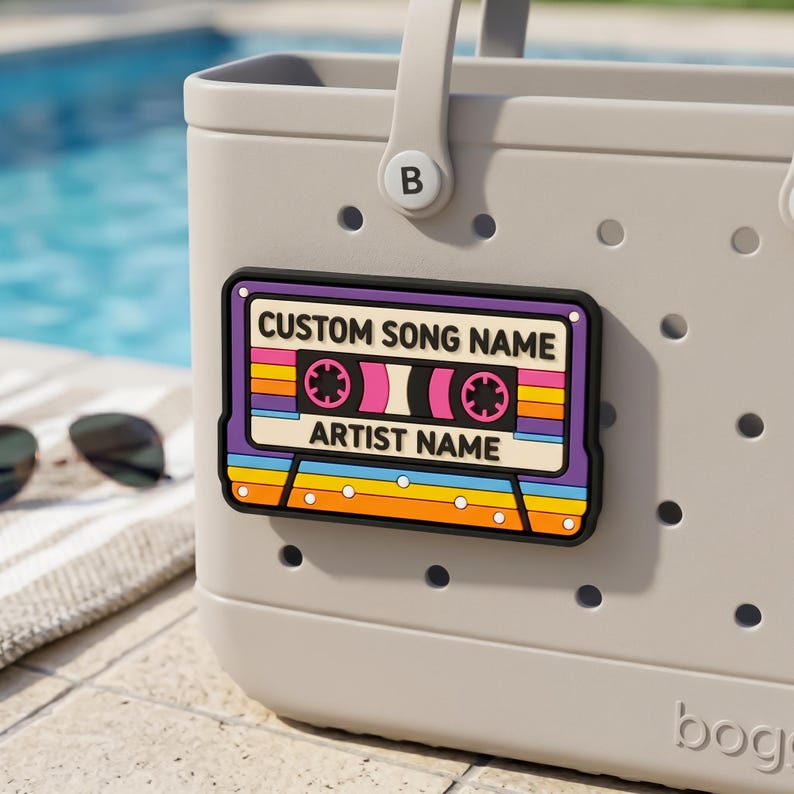 May include: A light gray tote bag with a colorful cassette tape design. The cassette tape has the text "CUSTOM SONG NAME" and "ARTIST NAME". The design features a black border, purple, pink, orange, yellow, and blue stripes.