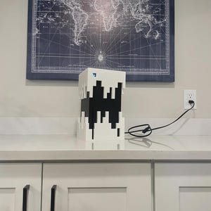 以下が含まれることがあります： ビデオゲームのキャラクターに似た、ピクセル化されたデザインの白と黒の幾何学的なランプ。 ランプはコンセントに差し込まれています。 世界地図が背景の壁に掛かっています。