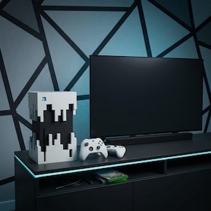 May include: A vertical gaming console featuring a black core encased in a white, pixelated, block-like shell with a blue Xbox logo. Two white wireless controllers with black buttons and joysticks sit next to the console in front of a flat screen TV.