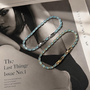 Op de afbeelding: Twee delicate armbanden met rechthoekige, lichtblauwe edelstenen. Eén armband heeft een zilveren sluiting, de andere een gouden sluiting. De armbanden worden getoond op een tijdschrift met de tekst "The Last Things Issue No.1".