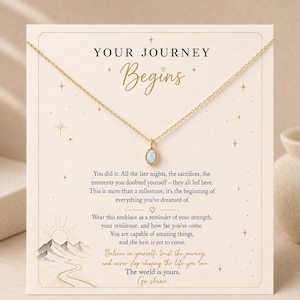 May include: A gold-colored necklace with a small, oval-shaped pendant. The pendant has a light blue and white stone. The necklace is displayed on a card with the words "YOUR JOURNEY Begins" and inspirational text.