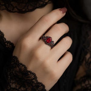 May include: A dark, gothic-style ring featuring a large, oval-shaped red gemstone as the centerpiece. The ring has an intricate, dark metalwork design with smaller red accents, creating a vintage and elegant aesthetic.