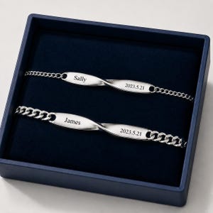Puede incluir: Dos pulseras plateadas con eslabones, presentadas en una caja azul oscuro. Cada pulsera presenta un nombre y una fecha grabados: "Sally" y "2023.5.21", y "James" y "2023.5.21". Las pulseras tienen un diseño retorcido.