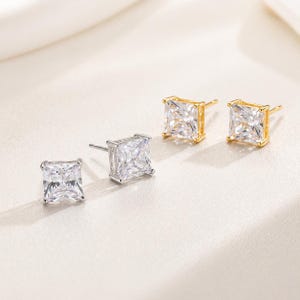 May include: Two pairs of square-cut stud earrings. One pair is silver-toned, and the other is gold-toned. Each earring features a clear, sparkling, square-shaped stone. The earrings are displayed on a light-colored surface.