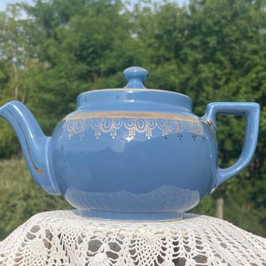 Peut inclure: Une théière en céramique bleu clair ornée d'une bande décorative dorée autour de la partie supérieure. La théière possède un couvercle assorti avec un petit bouton, un bec verseur incurvé et une anse rectangulaire, le tout avec des finitions dorées.