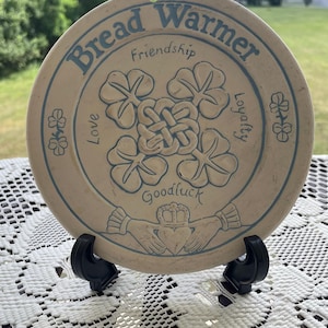 May include: A round, beige ceramic bread warmer featuring blue text and illustrations. The text reads Bread Warmer, Friendship, Love, Loyalty, and Goodluck. The design includes a central knot, four-leaf clovers, and two hands holding a heart.