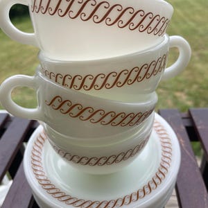 Può includere: Una pila di tazze da tè e piattini in vetro opalino bianco con un motivo a volute marrone ripetuto. Le tazze hanno manici arrotondati e i piattini sono circolari con un bordo decorativo marrone abbinato lungo il bordo esterno.
