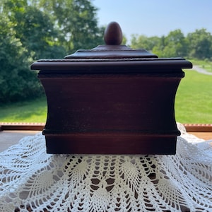 May include: A dark brown wooden box with a concave side profile and a lid featuring a central oval-shaped finial. The lid and base edges are decorated with a subtle, repeating embossed pattern, and the box has a smooth, matte finish.