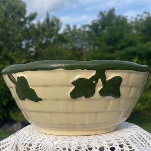 May include: A cream-colored ceramic bowl featuring a raised, dark green ivy leaf pattern around the rim and a textured basket-weave design on the exterior. The bowl has a smooth, glossy finish and a thick, dark green painted edge along the top.