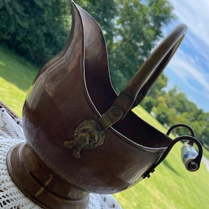 May include: A polished copper coal scuttle featuring a wide, curved opening, a sturdy arched handle, and decorative brass lion head mounts at the pivot points. A small, curved metal handle with a blue and white floral ceramic accent is attached.