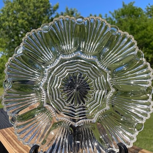 Könnte beinhalten: Ein Servierteller aus klarem Glas mit einem kreisförmigen Zentrum in strahlenförmigem Design, umgeben von einem breiten Rand aus zwölf ausgeprägten, muschelartigen, gewellten Abschnitten, die sich bis zum äußeren Rand des runden Tellers erstrecken.