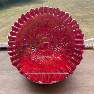 Puede incluir: Un plato redondo de vidrio de carnaval rojo con borde ondulado. El centro presenta un diseño en relieve de una herradura, tallos de trigo y flores tipo margarita. El texto "GOOD LUCK" aparece arqueado sobre la herradura con acabado iridiscente.