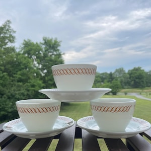 Puede incluir: Tres tazas de té de vidrio lechoso blanco con sus platos a juego, dispuestas en forma de pirámide. Cada taza y plato presenta un patrón repetitivo de volutas en forma de S de color naranja alrededor del borde. El conjunto es de vidrio blanco.
