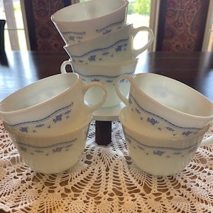 May include: A set of five white milk glass teacups featuring a delicate blue floral pattern around the center. Each cup has a rounded bowl shape, a small handle on the side, and a subtle ribbed texture along the lower exterior base of the vessel.
