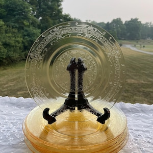 May include: A stack of translucent yellow glass plates featuring an intricate embossed floral pattern along the rim and a central medallion design. One plate is displayed upright on a black plastic stand to showcase the detailed texture.