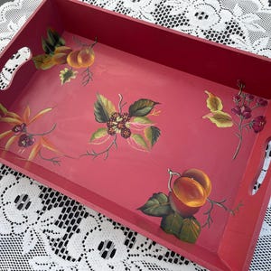 May include: A rectangular red wooden serving tray featuring hand-painted botanical illustrations of orange fruits, green leaves, and small dark berries. The tray includes two cutout handles on the shorter sides and a smooth, flat interior surface.