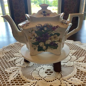 May include: A white ceramic teapot with a square-shaped body and a matching lid. The teapot features a colourful floral pattern with purple, yellow, and white flowers and green leaves. It has a curved spout, a handle, and four small feet.