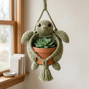 May include: A sage green crocheted turtle plant hanger featuring large dark eyes and a textured shell design. The turtle holds a terracotta pot containing a green succulent plant, suspended by a matching crocheted cord with a tassel at the base.