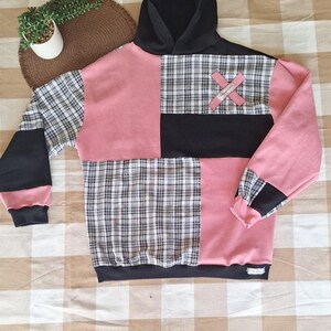 May include: A patchwork hoodie in pink, black, and plaid. The hoodie has a black hood, sleeves, and hem. The front is divided into sections of pink, black, and plaid fabric. An X-shaped patch is sewn on the plaid section.