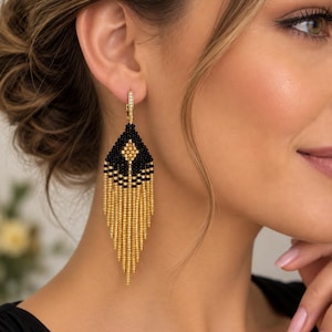 May include: A long, dangling earring featuring a diamond-shaped top section made of small black beads with a central gold bead accent. Below the diamond shape, numerous thin, vertical strands of metallic gold beads create a fringe effect.