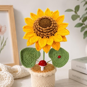 May include: A crochet sunflower with bright yellow petals and a textured brown centre, featuring two green leaves, a small red and white mushroom, and a tiny ladybug accent, all potted in a beige crocheted container with a scalloped top edge.