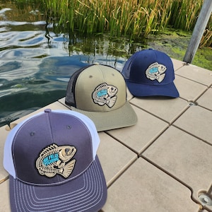 Könnte beinhalten: Drei Baseballkappen mit einem Fischlogo werden auf einem Steg gezeigt. Die Kappen sind marineblau, olivgrün und grau mit einem weißen Mesh-Rücken. Das Fischlogo trägt den Text "JOE FELIX FISHING" in Blau.
