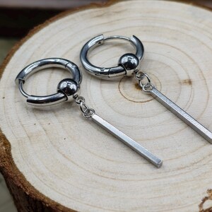 May include: A pair of silver-toned hoop earrings, each featuring a polished spherical bead on the hoop and a slender, rectangular bar pendant suspended from a small jump ring. The earrings have a smooth, metallic finish and a hinged closure.