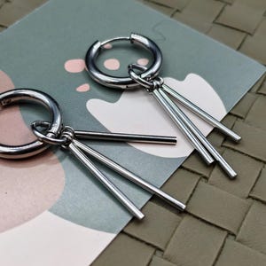 May include: A pair of silver-toned hoop earrings, each featuring three dangling thin metal bars attached to the bottom of the hoop. The earrings have a polished, reflective finish and a simple, clean design with a hinged closure mechanism.