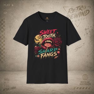 May include: A black short-sleeve t-shirt featuring a graphic print of a mouth with sharp fangs biting a lollipop. The design includes red roses, a yellow moon with bats, and the text Sweet Tooth, Sharp Fangs in red and teal lettering.