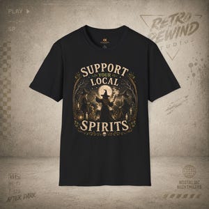 May include: A black short-sleeve t-shirt featuring a graphic print of three figures in pointed hats standing in a forest under a full moon. The text "SUPPORT YOUR LOCAL SPIRITS" is printed in a beige, arched font above and below the illustration.