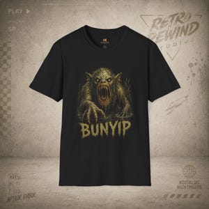 May include: A black short-sleeve t-shirt featuring a graphic print of a swamp creature with glowing eyes and sharp teeth emerging from water. The word BUNYIP is printed in a distressed, textured font below the creature in a muted gold color.