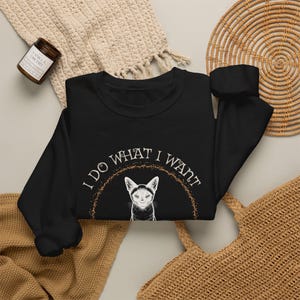 May include: A black long-sleeve sweatshirt featuring a white graphic of a cat centered below the text "I DO WHAT I WANT" arranged in a semi-circle. The text is framed by a speckled, arched design in a muted orange-brown color tone.