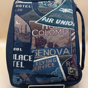 May include: A rectangular travel bag with a dark blue exterior and a tapestry design. The tapestry features travel-themed imagery, including the words "HOTEL", "COLOMB", "GENOVA", and "FLYING SERVICE". A handle is attached to the side.