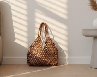 May include: A woven brown tote bag with a net-like design, featuring two handles. The bag is set against a white wall with shadows from blinds and rests on a light wood floor. The bag is empty.