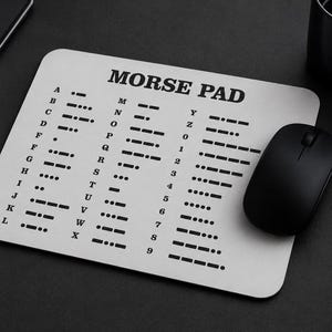 Puede incluir: Una alfombrilla de ratón rectangular blanca con el texto negro MORSE PAD en la parte superior, seguido de una lista completa del alfabeto y los números del cero al nueve, cada uno emparejado con su secuencia de código Morse de puntos y rayas.