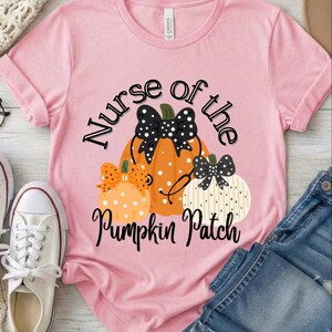 May include: A light pink short-sleeve t-shirt featuring a graphic of three pumpkins with polka dots and bows. The central orange pumpkin has a stethoscope draped over it. The text reads Nurse of the Pumpkin Patch in black cursive lettering.
