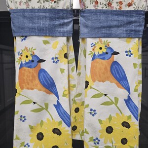 May include: Two kitchen towels with a bluebird and sunflower design. The towels have a white background with yellow sunflowers and blue accents. The top of the towels have a denim blue band and floral pattern.