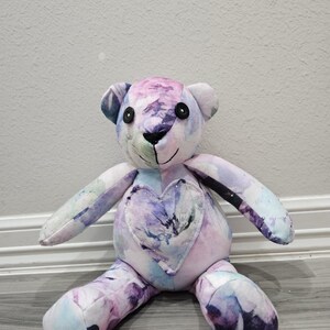 May include: A stuffed teddy bear crafted from fabric with a watercolor-style floral pattern in shades of purple, blue, and white. The bear features black button eyes, a stitched nose and mouth, and a heart-shaped patch on its chest.