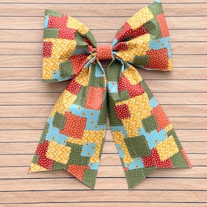 May include: A large fabric hair bow featuring a patchwork design of various squares in red, yellow, light blue, and olive green. The patterns include small white flowers, white polka dots, and solid colors, all tied together with a central orange knot.