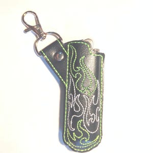 May include: Black leather lighter case with a green and white embroidered flame design. The case has a silver metal clip for attaching to a keychain.